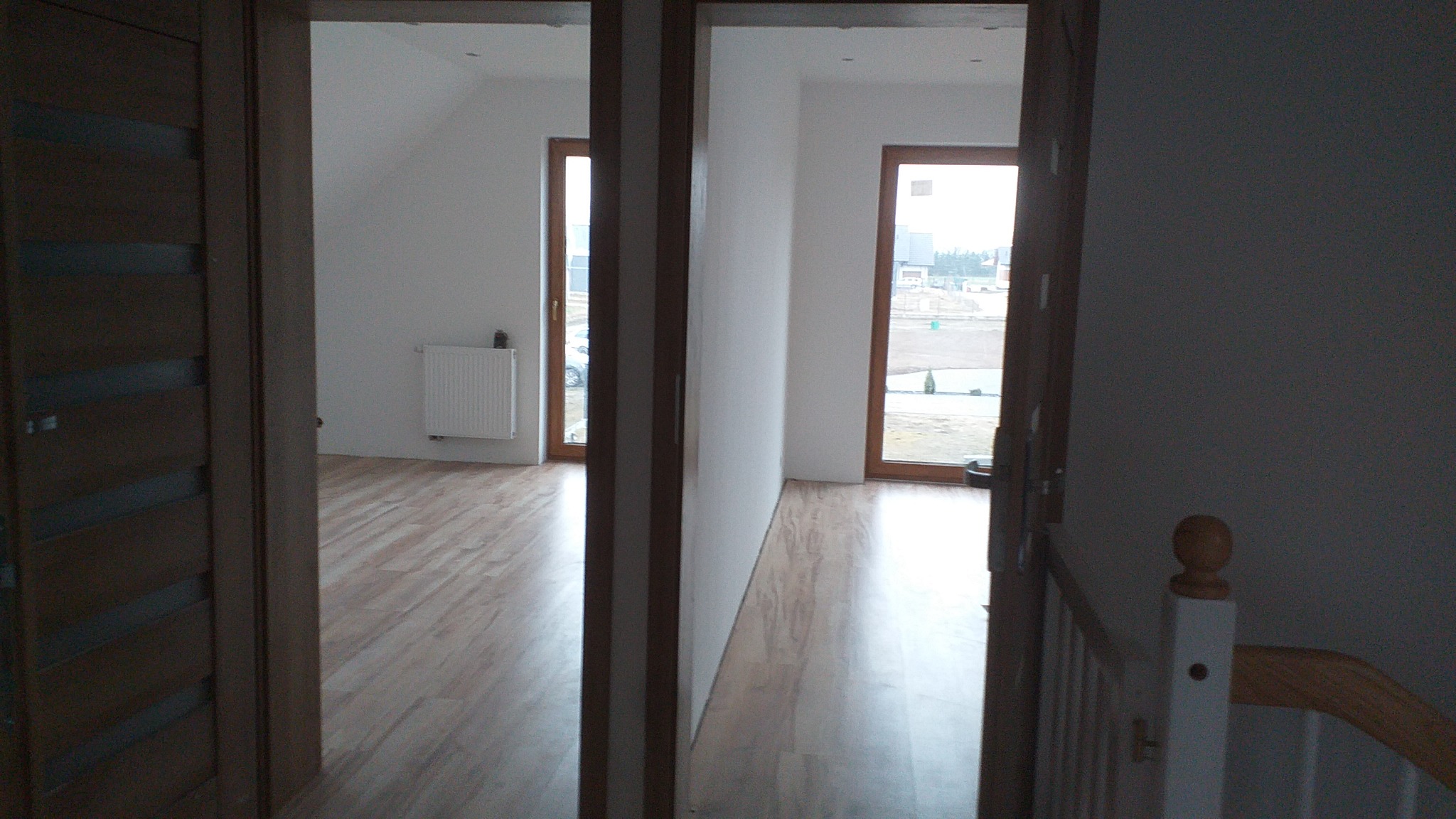 Widok przez dwa otwory drzwiowe na świeżo wykończone wnętrze w Złotowie: jasne ściany, drewniana podłoga, widoczne okna i grzejnik.
