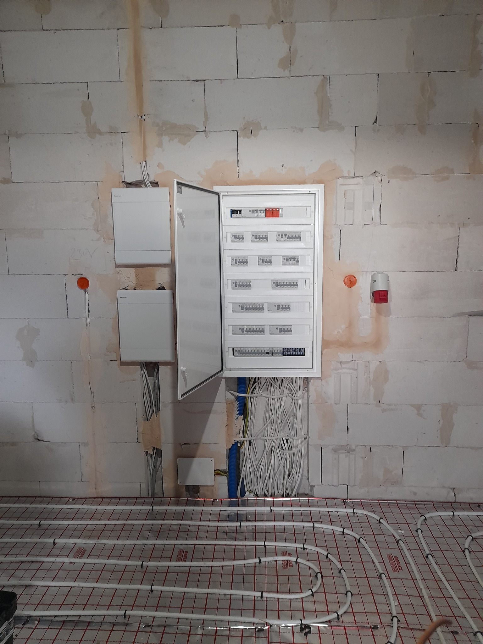Otwarta skrzynka rozdzielcza z licznymi kablami na surowej ścianie z betonu komórkowego, widoczna instalacja ogrzewania podłogowego.