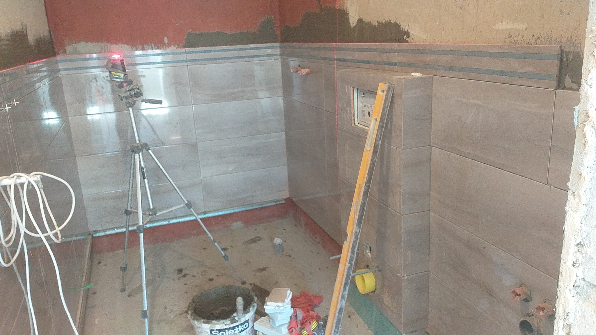 Układanie płytek w łazience z użyciem poziomicy laserowej na statywie, widoczny kubeł z klejem Spieźko, poziomica i fragment niezakończonej ściany.