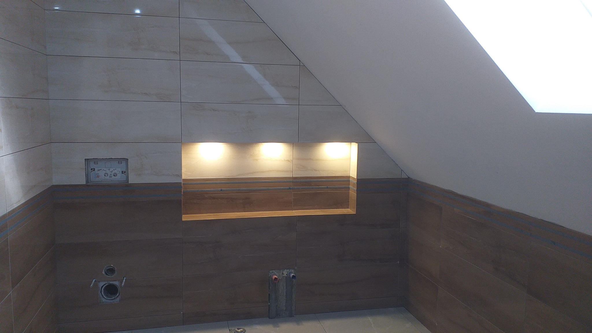 Łazienka w trakcie prac wykończeniowych na poddaszu, widoczne płytki w odcieniach beżu i brązu, wnęka z oświetleniem, przygotowane instalacje wodno-kanalizacyjne, oraz otwór na stelaż podtynkowy WC.