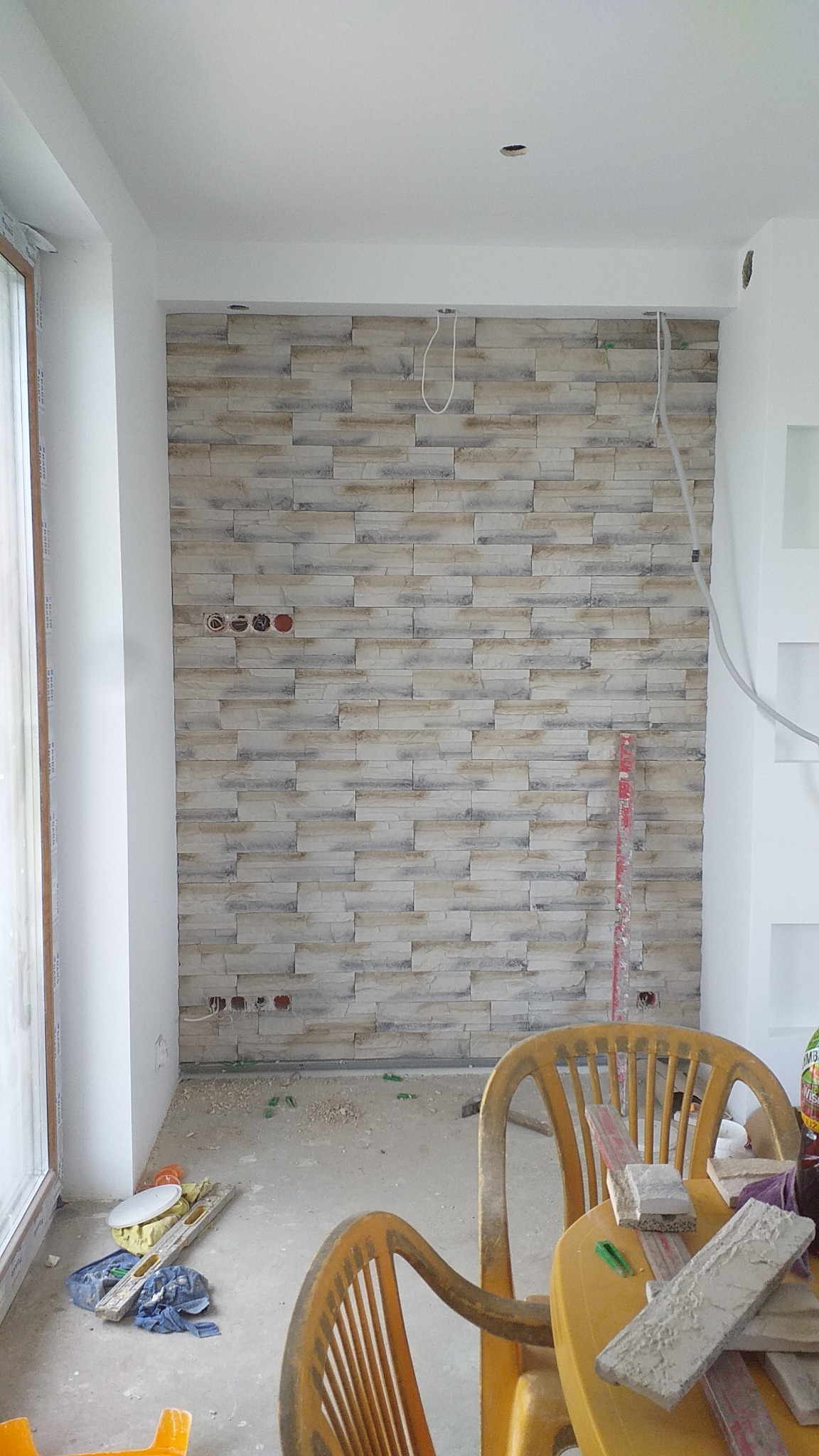 Ściana wyłożona dekoracyjną cegłą w trakcie wykończenia, widoczne puszki elektryczne, przewody, poziomica i narzędzia na podłodze.
