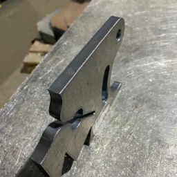 Precyzyjnie wycięty, surowy element stalowy z otworem i charakterystycznym kształtem, wyprodukowany w Inowrocławiu, leży na metalowym stole roboczym.