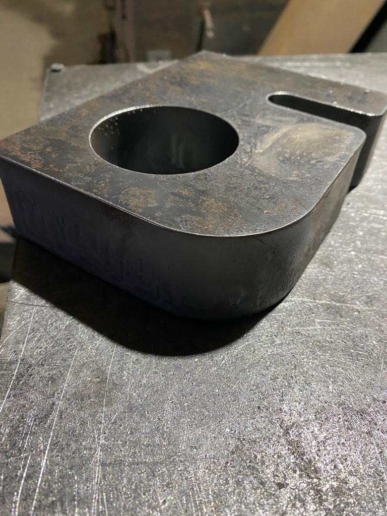 Surowy, gruby element stalowy z otworem i wycięciem, leżący na metalowym stole warsztatowym z widocznymi śladami obróbki.