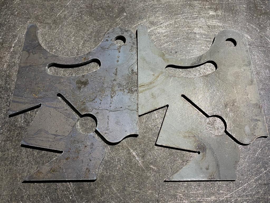 Dwie wycięte laserowo, stalowe blachy z otworami i wycięciami, leżące na metalowym stole warsztatowym, widoczne ślady obróbki.