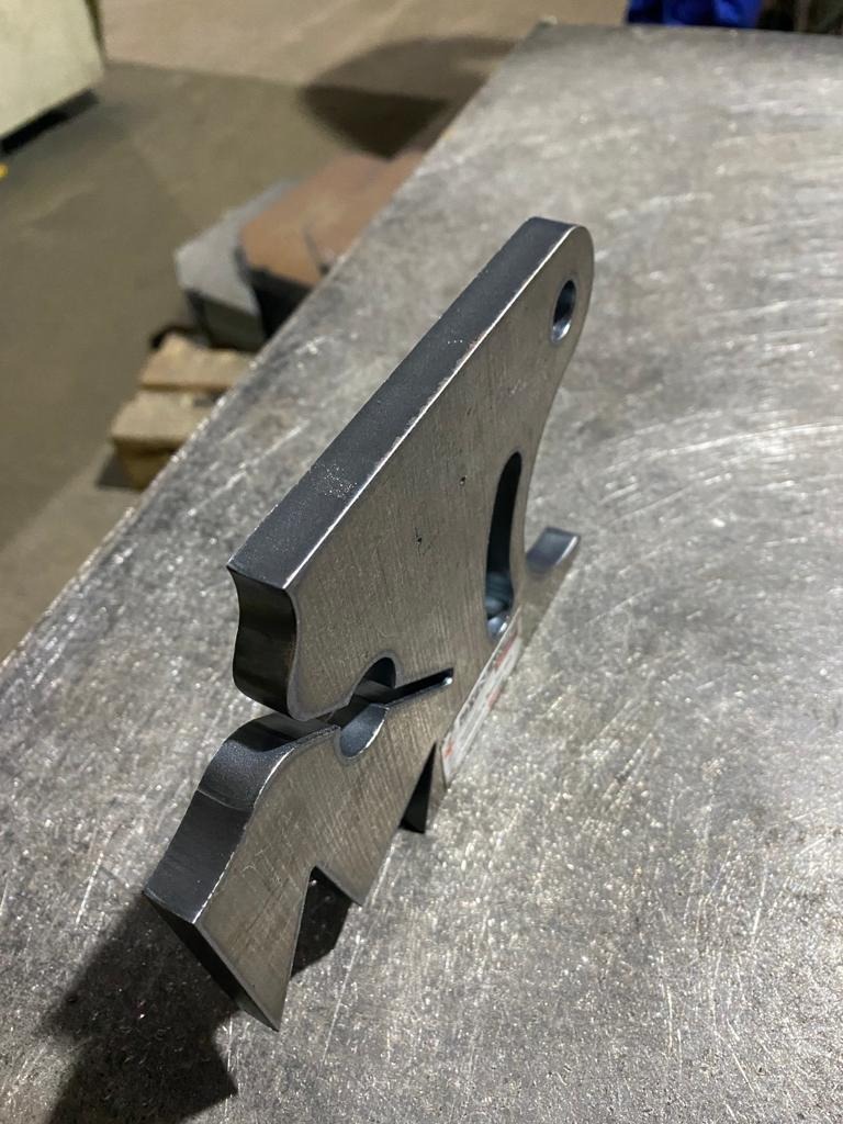 Precyzyjnie wycięty, surowy element stalowy z otworem i charakterystycznym kształtem, wyprodukowany w Inowrocławiu, leży na metalowym stole roboczym.