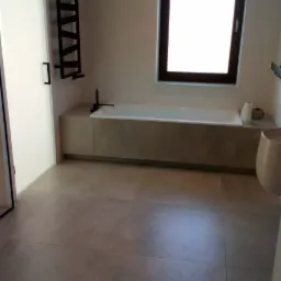 Nowoczesna łazienka z dużą wanną obudowaną płytkami w odcieniach szarości, czarnym grzejnikiem drabinkowym i oknem z ciemną ramą.