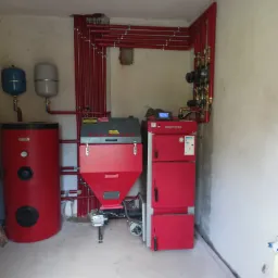 Czerwony piec Defro z zasobnikiem i zbiornikiem buforowym, orurowanie grzewcze w kolorze czerwonym, zainstalowane w kotłowni z surowymi ścianami.