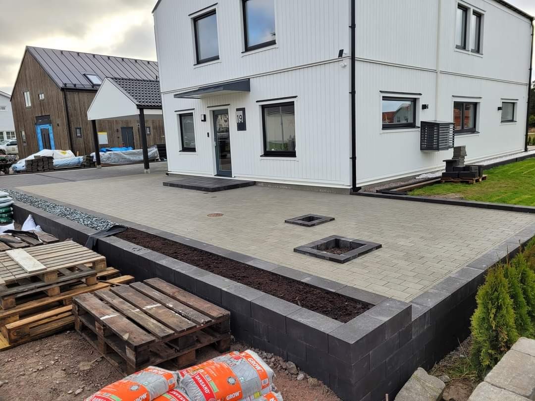 Nowoczesna posesja z jasnoszarą kostką brukową, obrzeżami z ciemnej kostki, podwyższonymi rabatami i paletami z workami ziemi w tle.