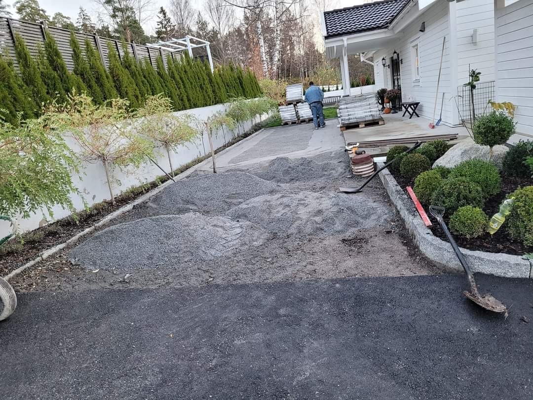 Przygotowanie podłoża pod kostkę brukową na podjeździe do białego domu z zielonymi krzewami i drzewkami, widoczne narzędzia i palety z kostką.