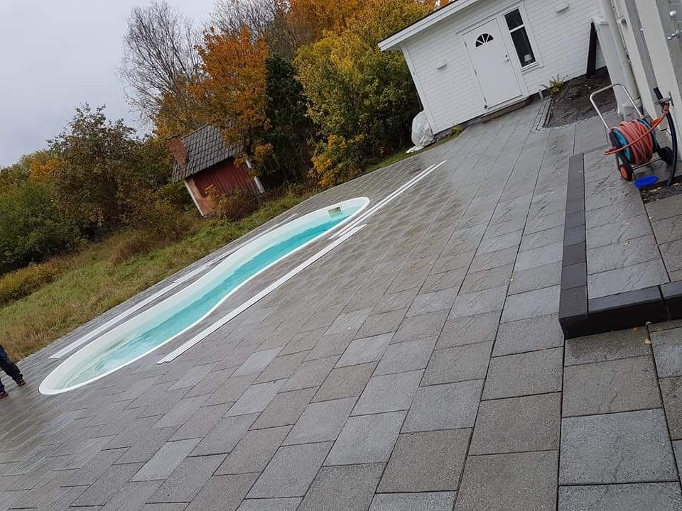 Ułożona szara kostka brukowa wokół owalnego basenu, widoczny fragment budynku gospodarczego i domek na wzgórzu w tle.