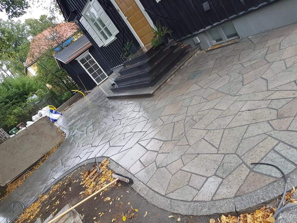 Świeżo ułożona nieregularna kostka na podjeździe domu z czarną elewacją, widoczne liście i szczotka brukarska.