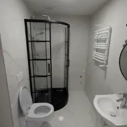 Nowoczesna łazienka z kabiną prysznicową w czarnej ramie, białą umywalką z szafką, toaletą z otwartą klapą i grzejnikiem drabinkowym, wykończona białymi płytkami imitującymi marmur.