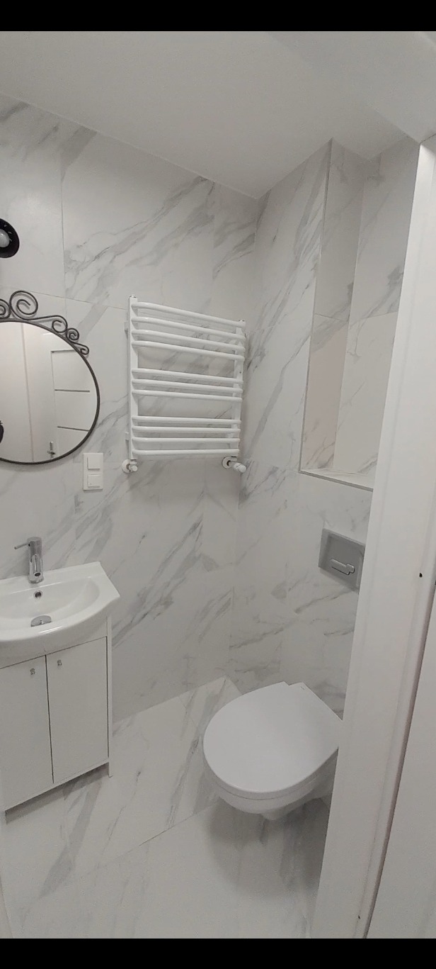 Elegancka, wykończona łazienka w bieli i szarości z marmurkowym wzorem na płytkach, wyposażona w umywalkę z szafką, ozdobne lustro, grzejnik drabinkowy i podwieszaną toaletę.