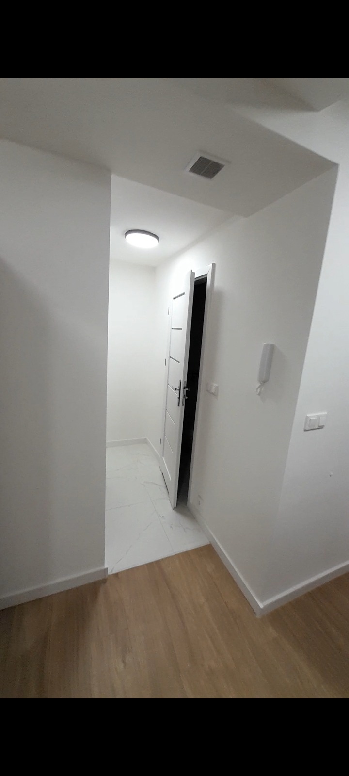 Wnętrze korytarza w Łodzi po remoncie: białe ściany, drewnopodobna podłoga przechodząca w białe płytki w drugim pomieszczeniu, białe drzwi z klamką i oświetlenie sufitowe.