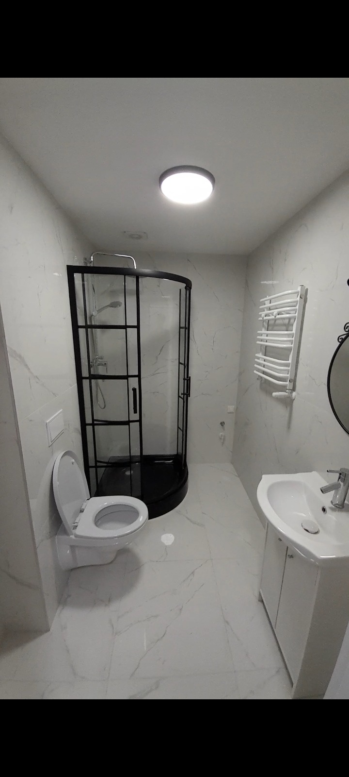 Nowoczesna łazienka z kabiną prysznicową w czarnej ramie, białą umywalką z szafką, toaletą z otwartą klapą i grzejnikiem drabinkowym, wykończona białymi płytkami imitującymi marmur.