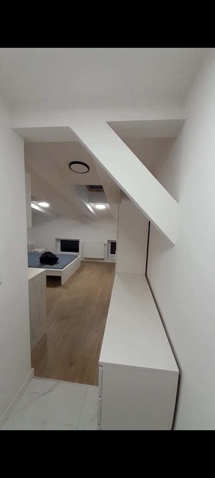 Nowoczesne poddasze ze skosami i zabudową meblową w bieli, widok z korytarza na pokój z drewnianą podłogą i łóżkiem.
