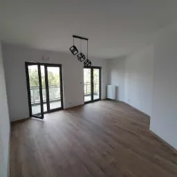 Jasne, przestronne wnętrze po remoncie z parkietem w odcieniach brązu, białymi ścianami, nowoczesną lampą wiszącą i dwoma balkonami z czarnymi ramami okiennymi.