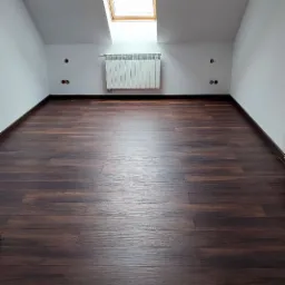 Poddasze z oknem dachowym, grzejnikiem i świeżo ułożonymi, ciemnymi panelami podłogowymi z widoczną strukturą drewna.