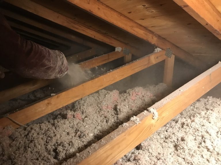 Aplikacja izolacji celulozowej w przestrzeni poddasza, widoczny wąż i rozpylana izolacja pomiędzy drewnianymi belkami stropowymi, częściowo wypełniona przestrzeń.