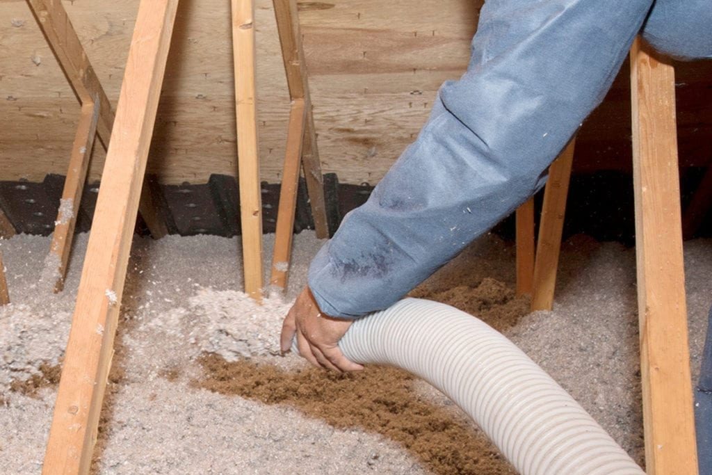 Pracownik w kombinezonie aplikuje izolację z rury do przestrzeni poddasza między drewnianymi krokwiami, widoczne włókna izolacyjne w kolorze szarym i brązowym.