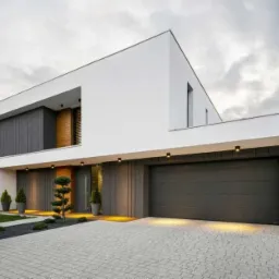 Nowoczesny, dwukondygnacyjny dom jednorodzinny z płaskim dachem, szarą bramą garażową i elewacją w odcieniach bieli i szarości, oświetlony punktowymi lampami, z kostką brukową na podjeździe...