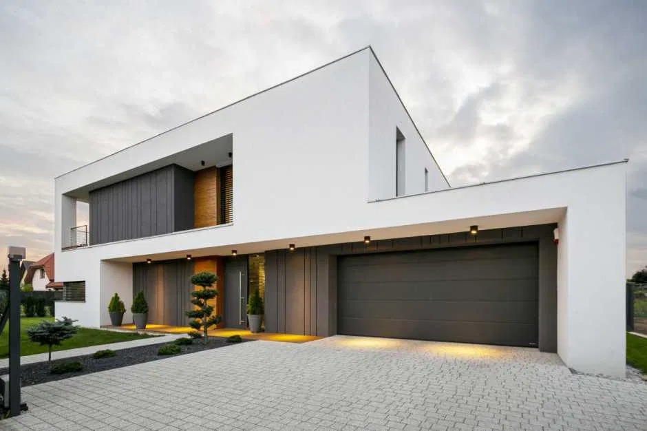 Nowoczesny, dwukondygnacyjny dom jednorodzinny z płaskim dachem, szarą bramą garażową i elewacją w odcieniach bieli i szarości, oświetlony punktowymi lampami, z kostką brukową na podjeździe...