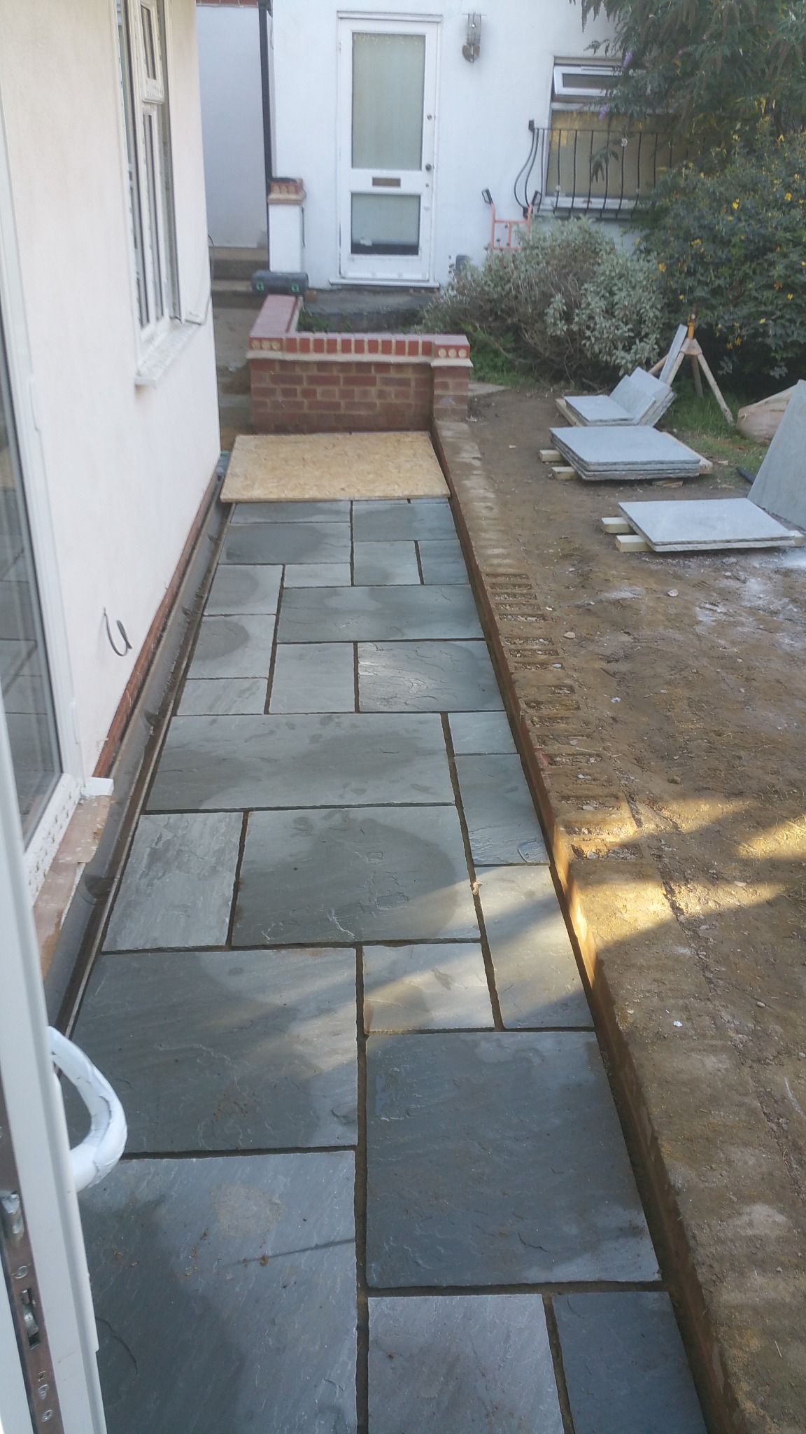 Układanie szarej kamiennej ścieżki prowadzącej do wejścia domu, widoczne płyty kamienne, krawężnik z cegły i przygotowany grunt