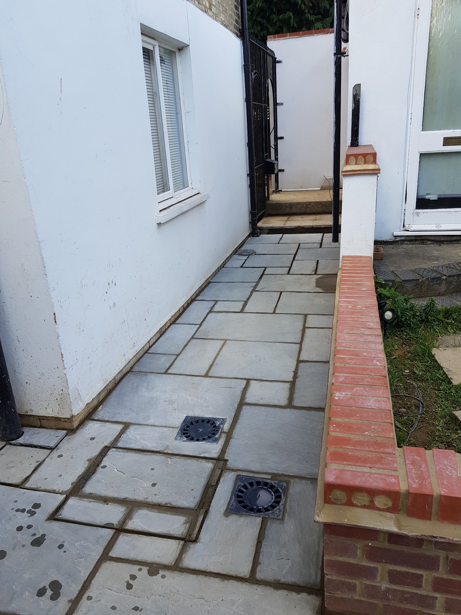 Ułożona szara kostka brukowa na ścieżce obok białego budynku z oknem, z czarną kratką kanalizacyjną i niskim murkiem z czerwonej cegły po prawej stronie.