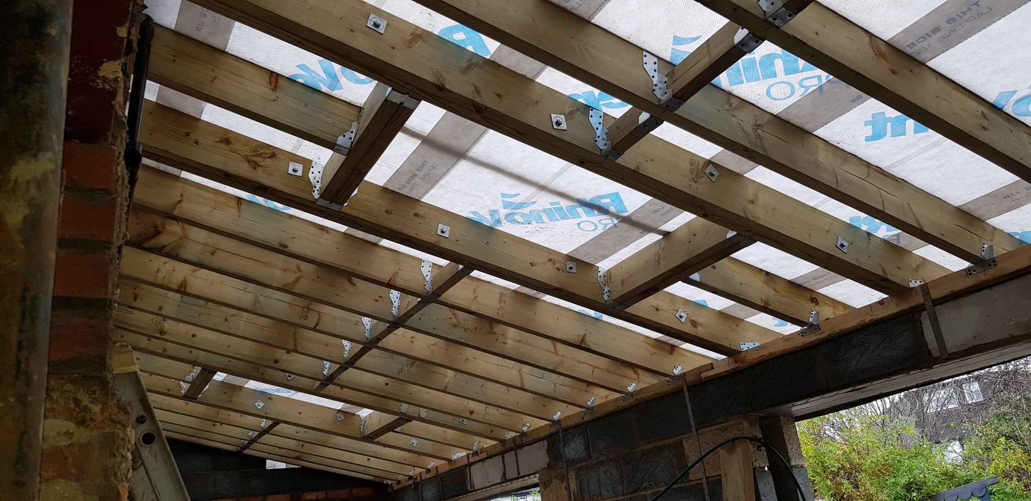 Konstrukcja dachu z drewnianych belek, zabezpieczona membraną z widocznym nadrukiem producenta, mocowana metalowymi łącznikami do muru z bloczków betonowych.