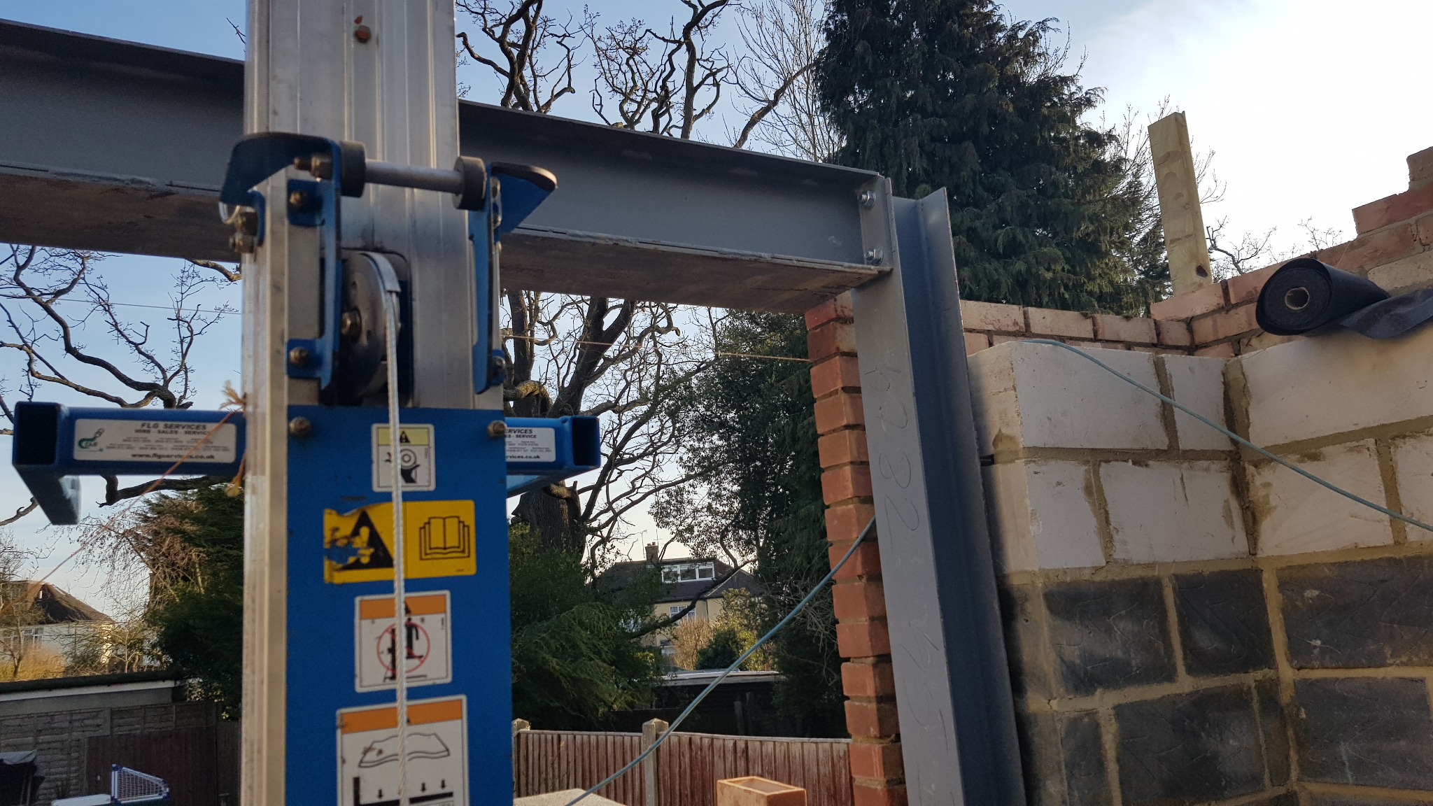 Montaż stalowej belki nad otworem w murowanej ścianie, widoczny podnośnik z niebieską obudową i naklejkami ostrzegawczymi, w tle drzewa i budynek mieszkalny.