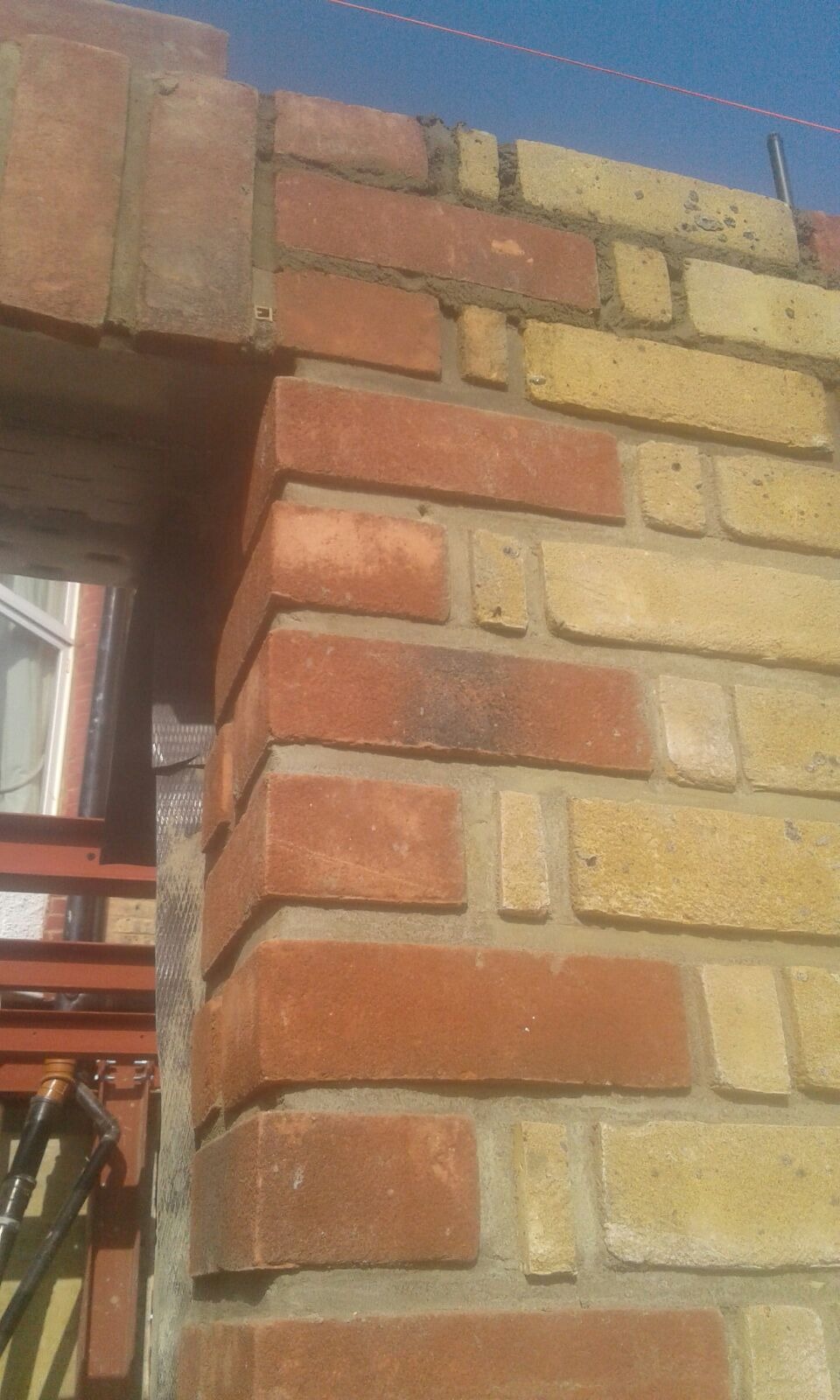 Fragment muru z cegły klinkierowej w odcieniach czerwieni i żółci, z widocznym oknem w tle i elementami rusztowania.