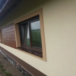 Elewacja domu z oknem w brązowej ramie i fragmentem ściany z fakturą drewna, wykończona w jasnych beżach.