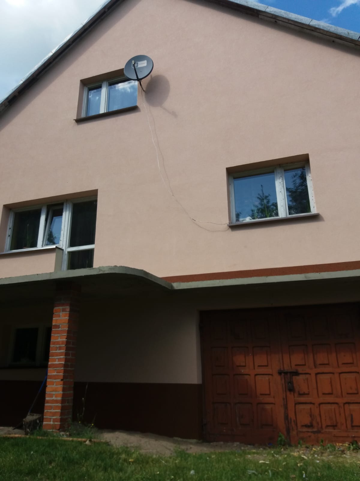 Dwukondygnacyjny dom z elewacją w odcieniach beżu, z widoczną anteną satelitarną na górnej kondygnacji, okna z białymi ramami i brązowe drzwi garażowe na poziomie gruntu.