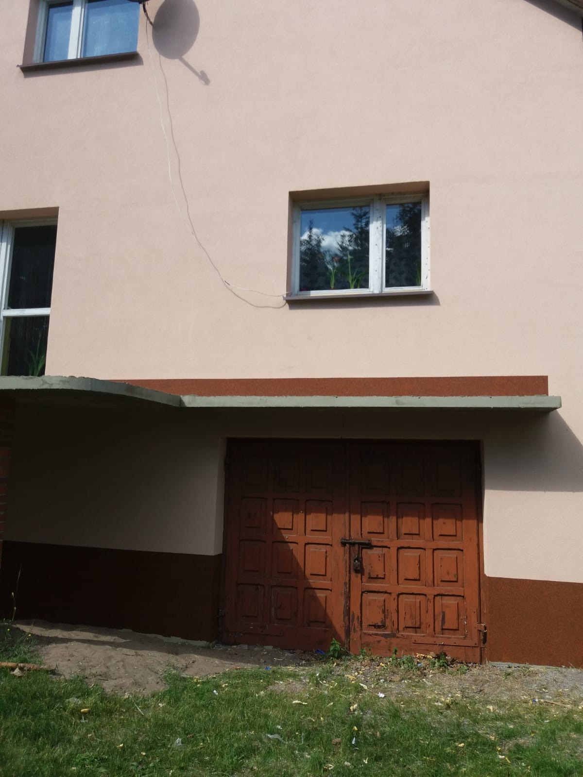 Dwukondygnacyjny budynek w Radomiu z ocieploną elewacją w kolorze beżowym, brązowe detale wykończeniowe, widoczne okna z kwiatami i brązowa brama garażowa z wyraźnymi śladami użytkowania.