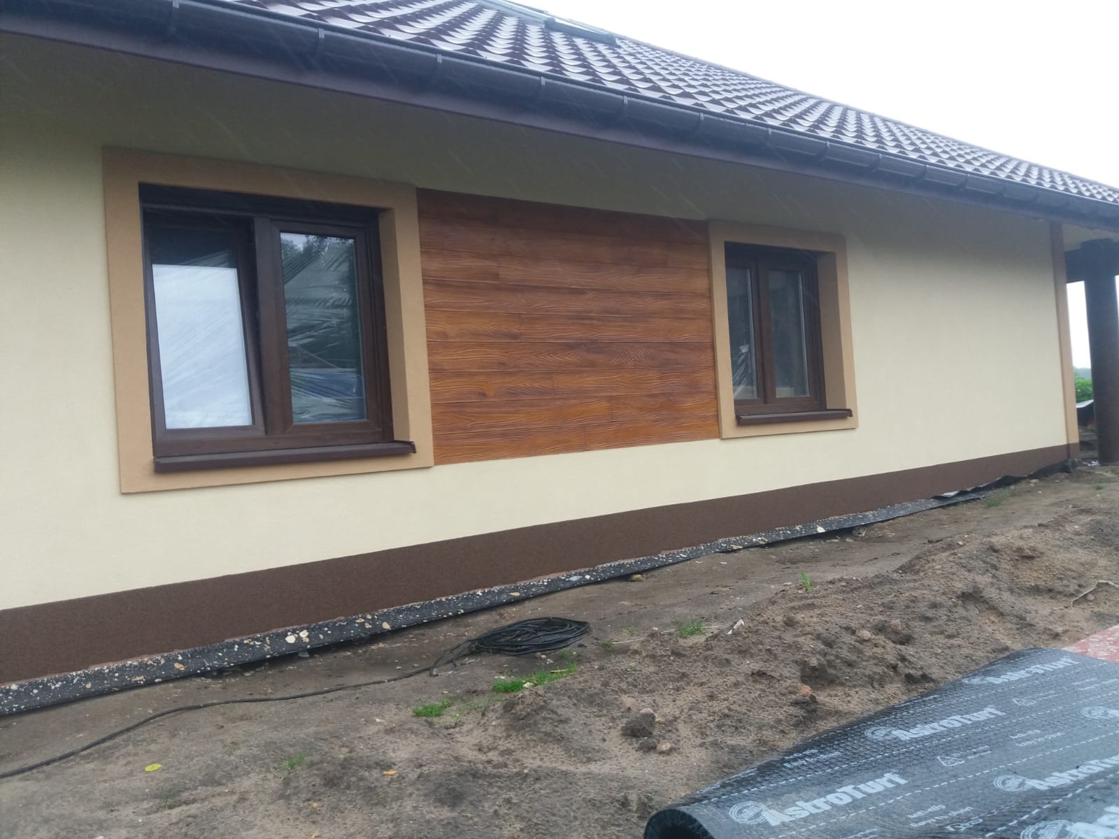 Fragment elewacji domu jednorodzinnego z dekoracyjnym panelem imitującym drewno, brązowe okna z zabezpieczeniem folią oraz przygotowany grunt pod ocieplenie.