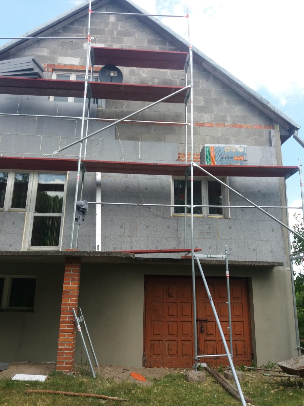 Dom w trakcie prac: częściowo ocieplona szara elewacja z widocznymi rusztowaniami, paczka materiału na rusztowaniu, ceglana kolumna i brązowe, drewniane drzwi garażowe.