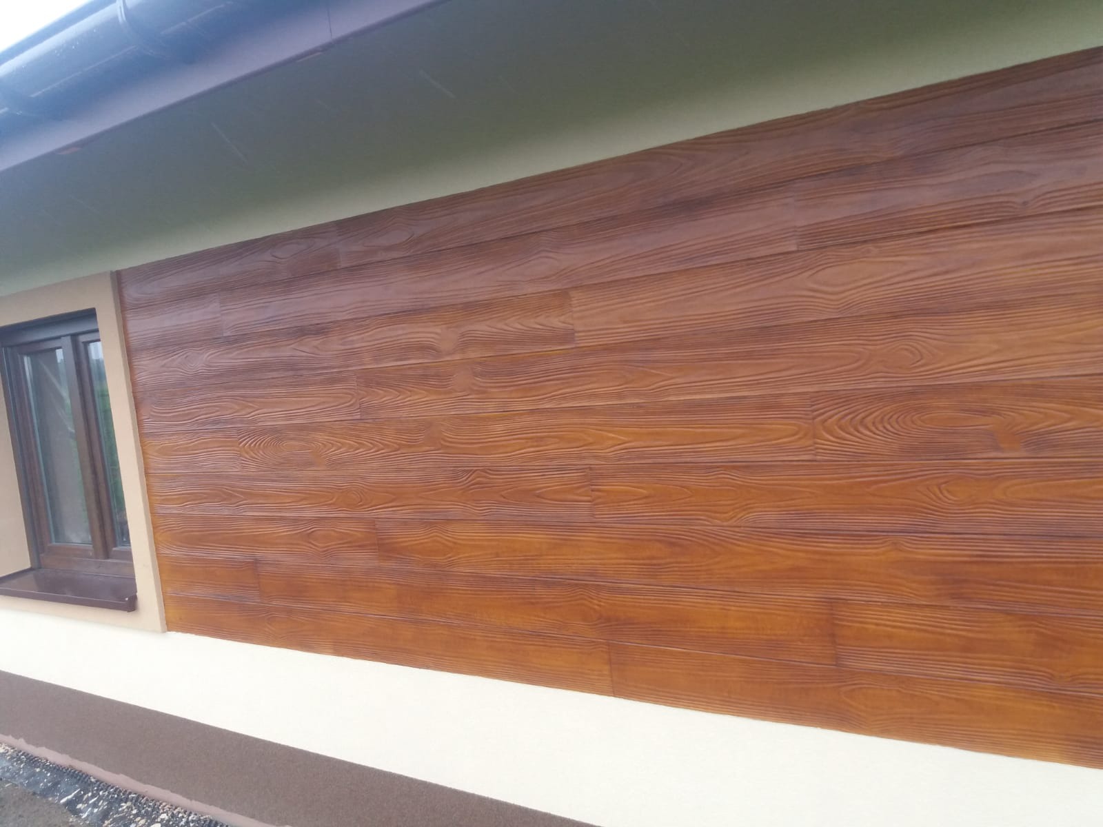 Fragment ściany domu z brązową elewacją imitującą drewno, widoczny fragment okna i podbitki dachowej.