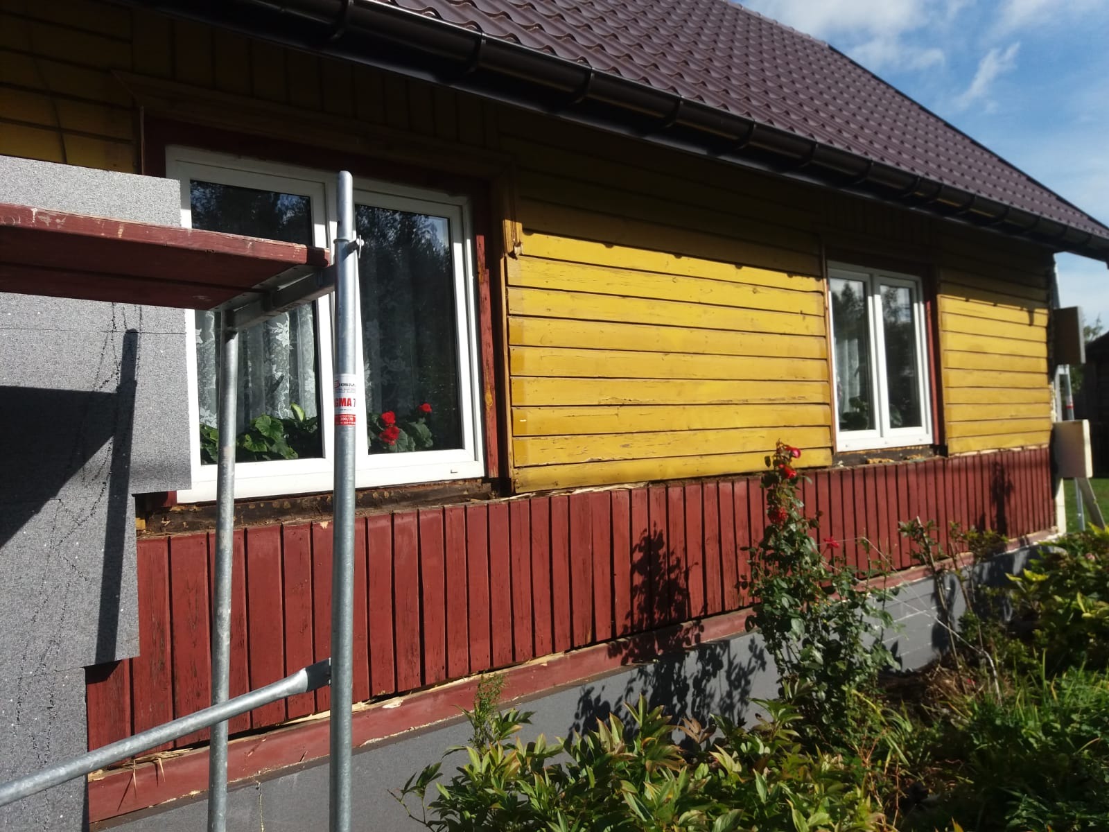 Fragment żółtego, drewnianego domu z widocznym ociepleniem styropianem przy oknie, czerwona podmurówka i rusztowanie, w tle róże w oknie i przed domem.