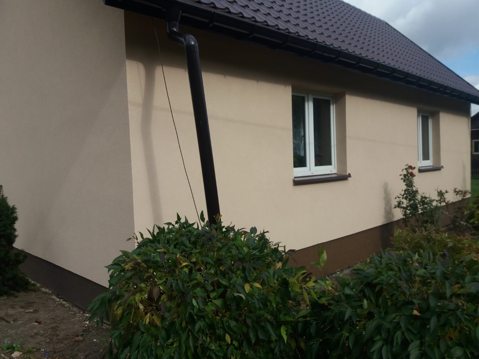 Dom jednorodzinny z beżową elewacją, brązowym dachem i rynną, widoczne krzewy w ogrodzie.