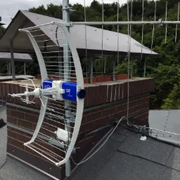 Antena telewizyjna typu Yagi-Uda, zamontowana na kominie z cegły, z widocznym kablem koncentrycznym i otaczającą zielenią.