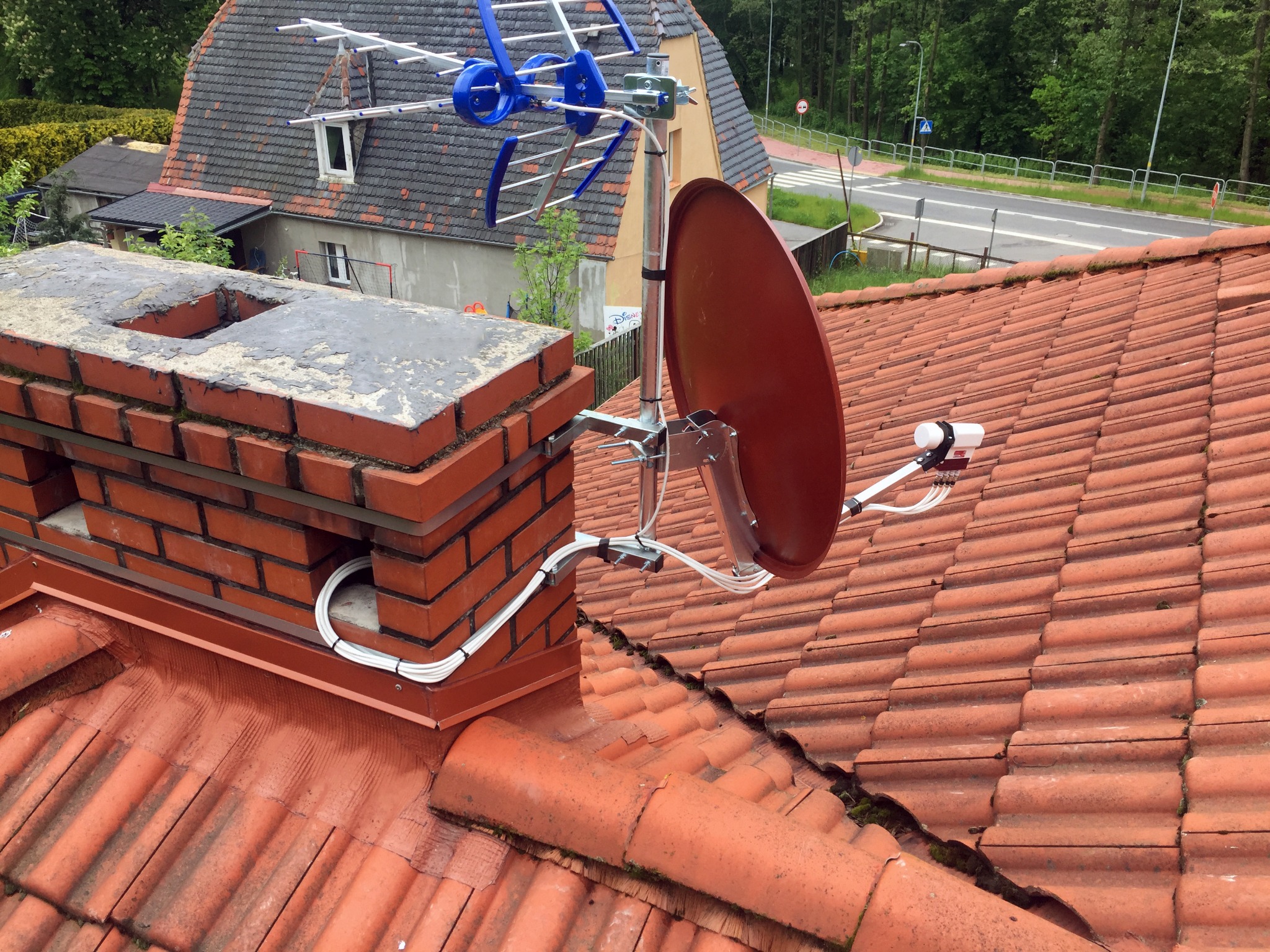 Antena satelitarna i antena telewizyjna zamontowane na dachu z czerwonej dachówki, przymocowane do komina z cegły, widoczne przewody antenowe.