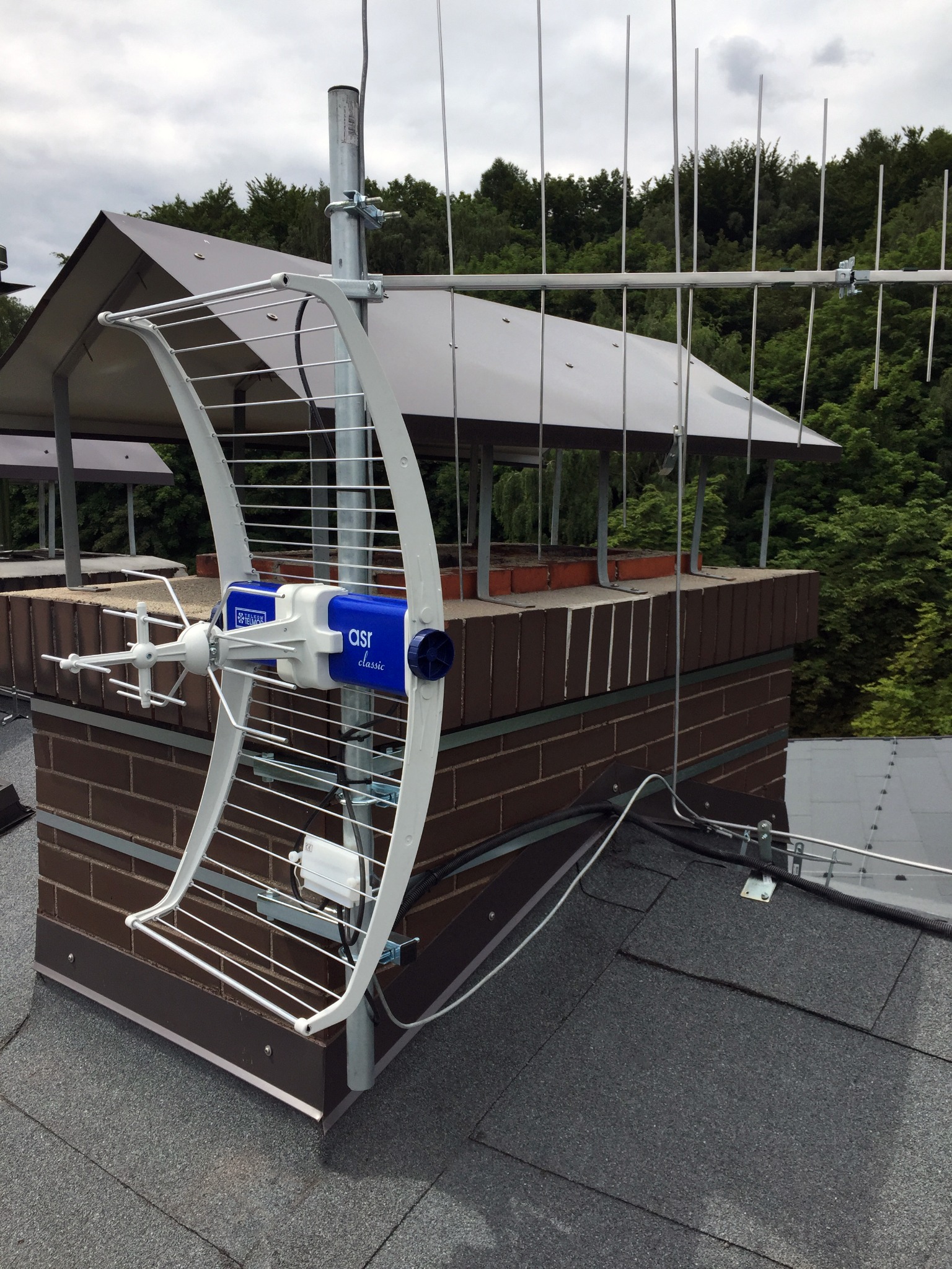 Antena telewizyjna typu Yagi-Uda, zamontowana na kominie z cegły, z widocznym kablem koncentrycznym i otaczającą zielenią.