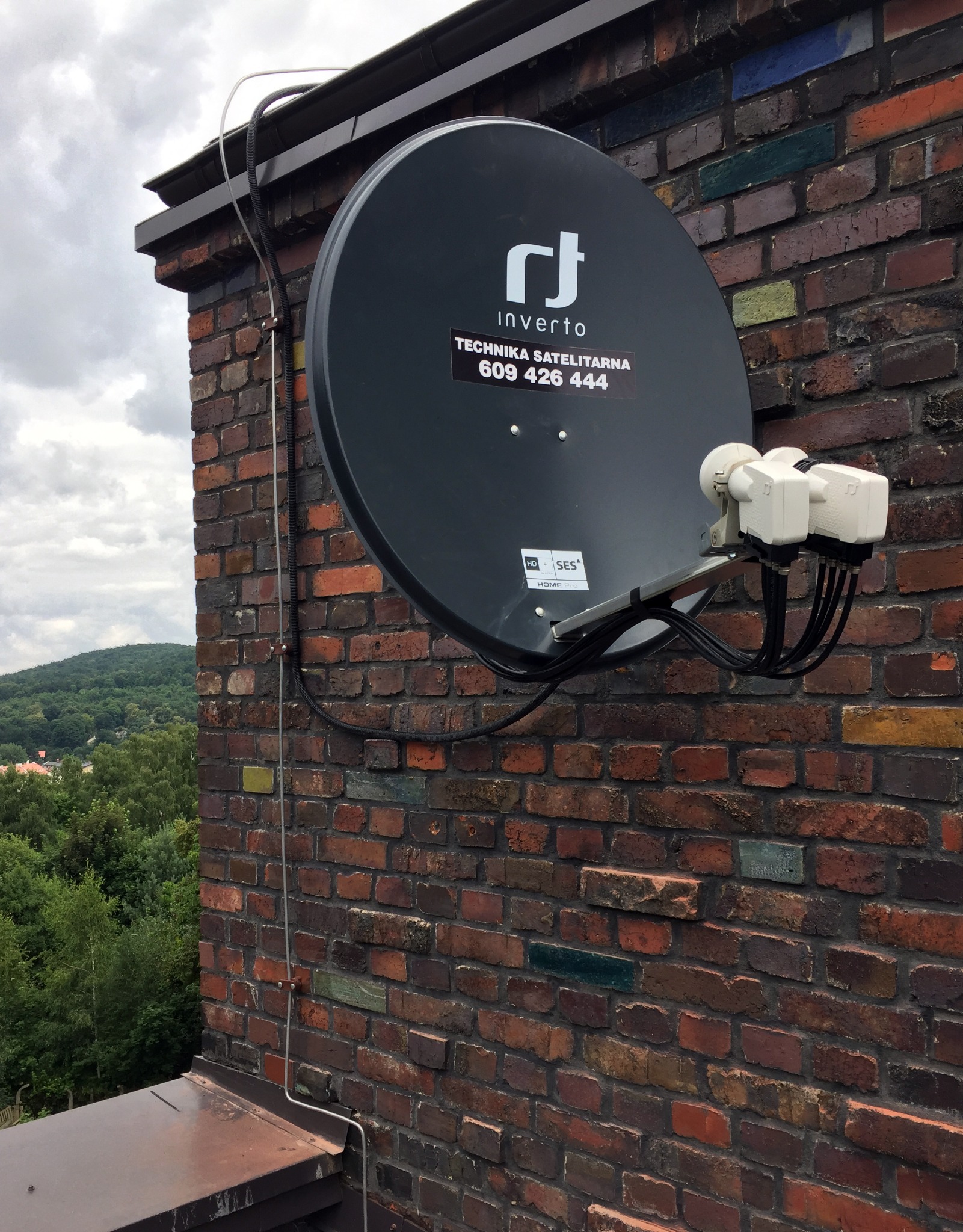 Czarna antena satelitarna Invertro zamontowana na starej, wielobarwnej ceglanej ścianie budynku, z widocznymi przewodami i elementami mocującymi. W tle zieleń drzew i zabudowania.