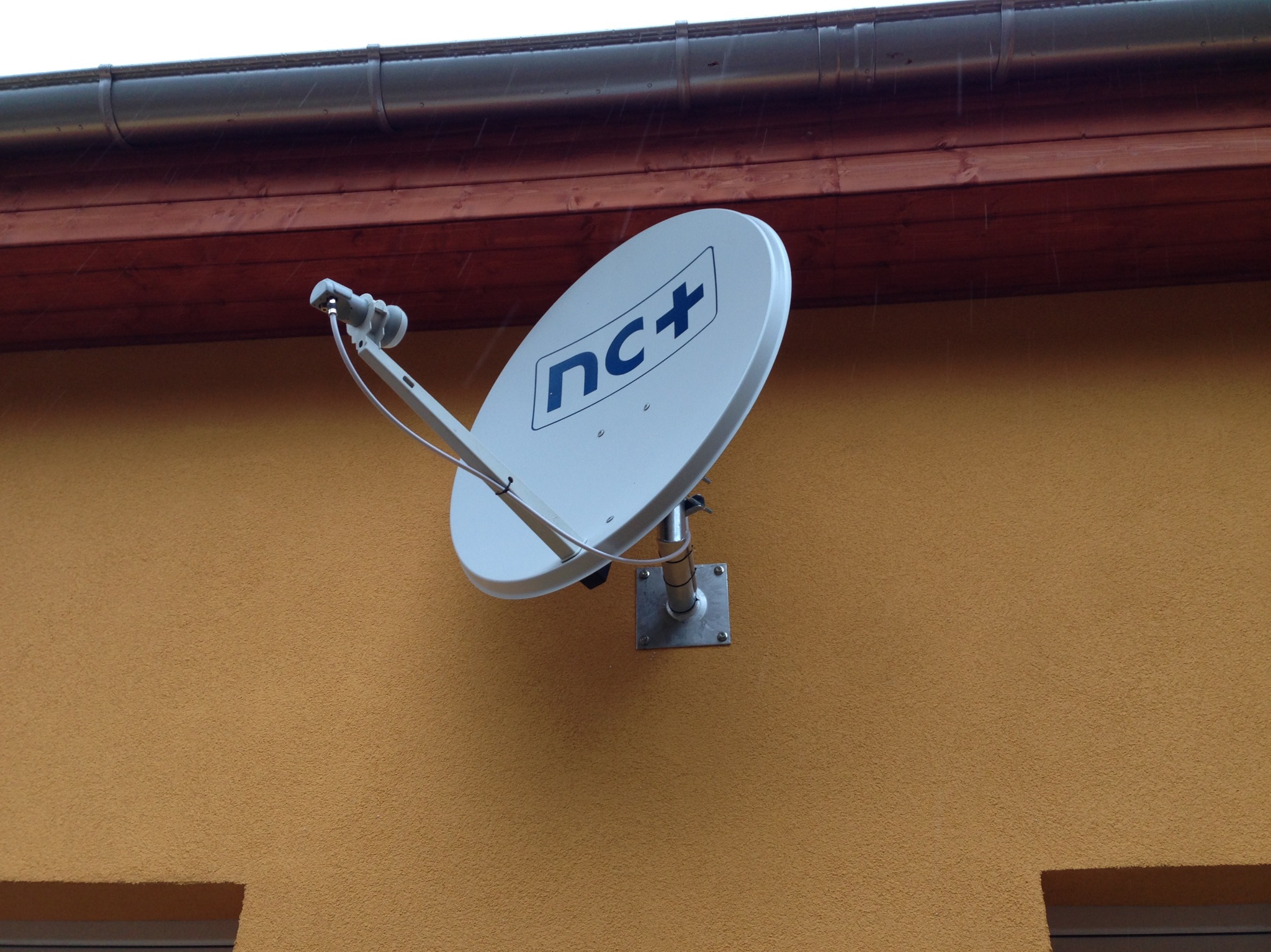 Biała antena satelitarna z logo nc+ zamontowana na pomarańczowej ścianie budynku pod brązowym dachem, widoczne krople deszczu.