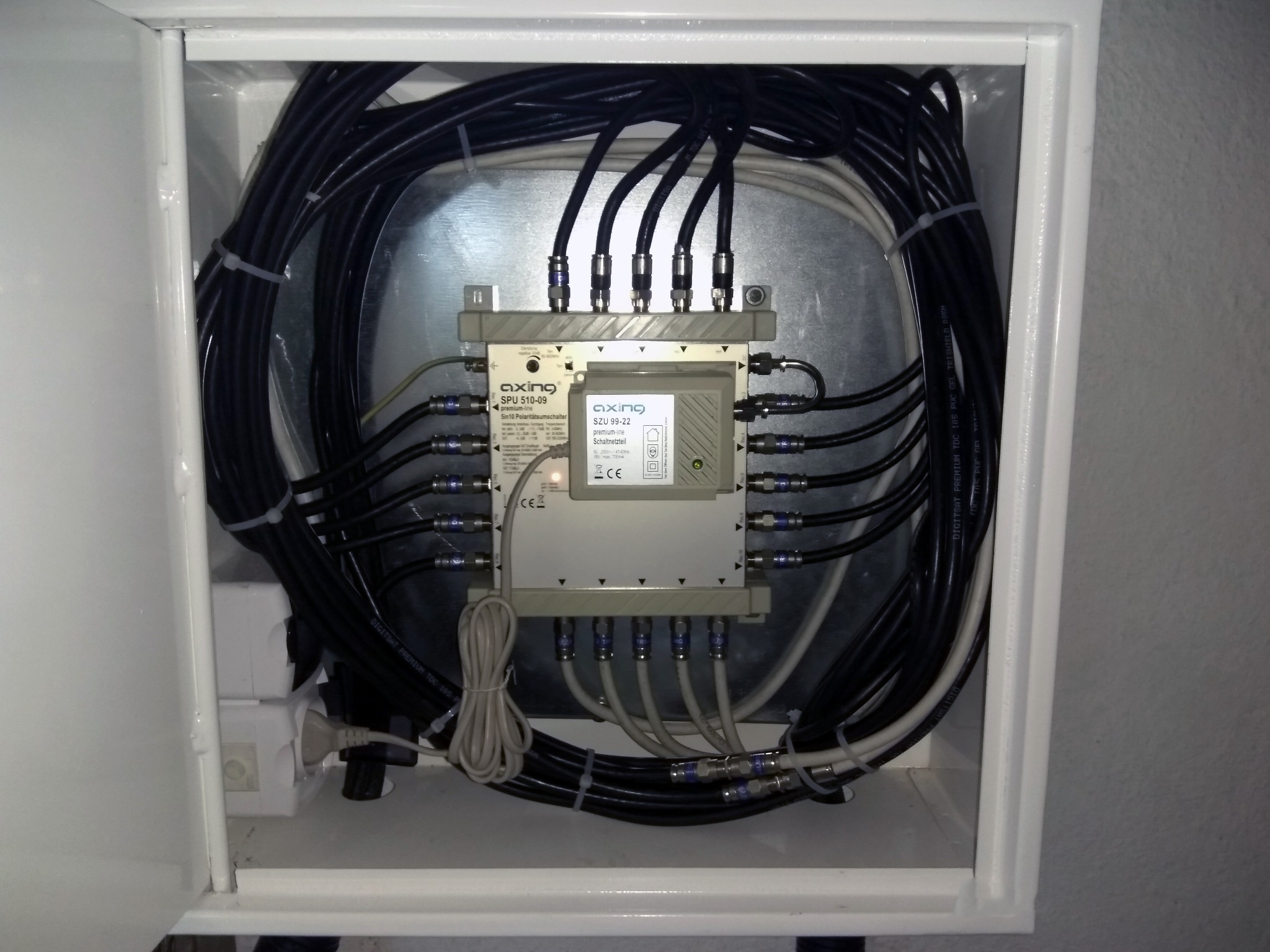 Rozdzielacz sygnału telewizyjnego Axing SPU 510-09 i zasilacz SZU 99-22 w białej skrzynce, z podłączonymi kablami koncentrycznymi i zasilaniem sieciowym.