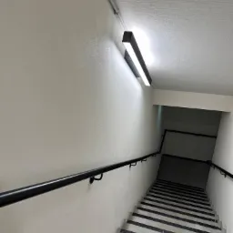 Klatka schodowa z szarymi ścianami, czarną poręczą i paskami antypoślizgowymi na schodach, oświetlona podłużną lampą sufitową.