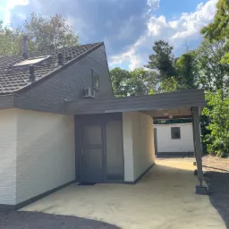 Odnowiona elewacja domu jednorodzinnego z jasną cegłą, szarą stolarką okienną i drzwiową oraz zadaszeniem nad wejściem, widoczny fragment ogrodu i błękitne niebo.