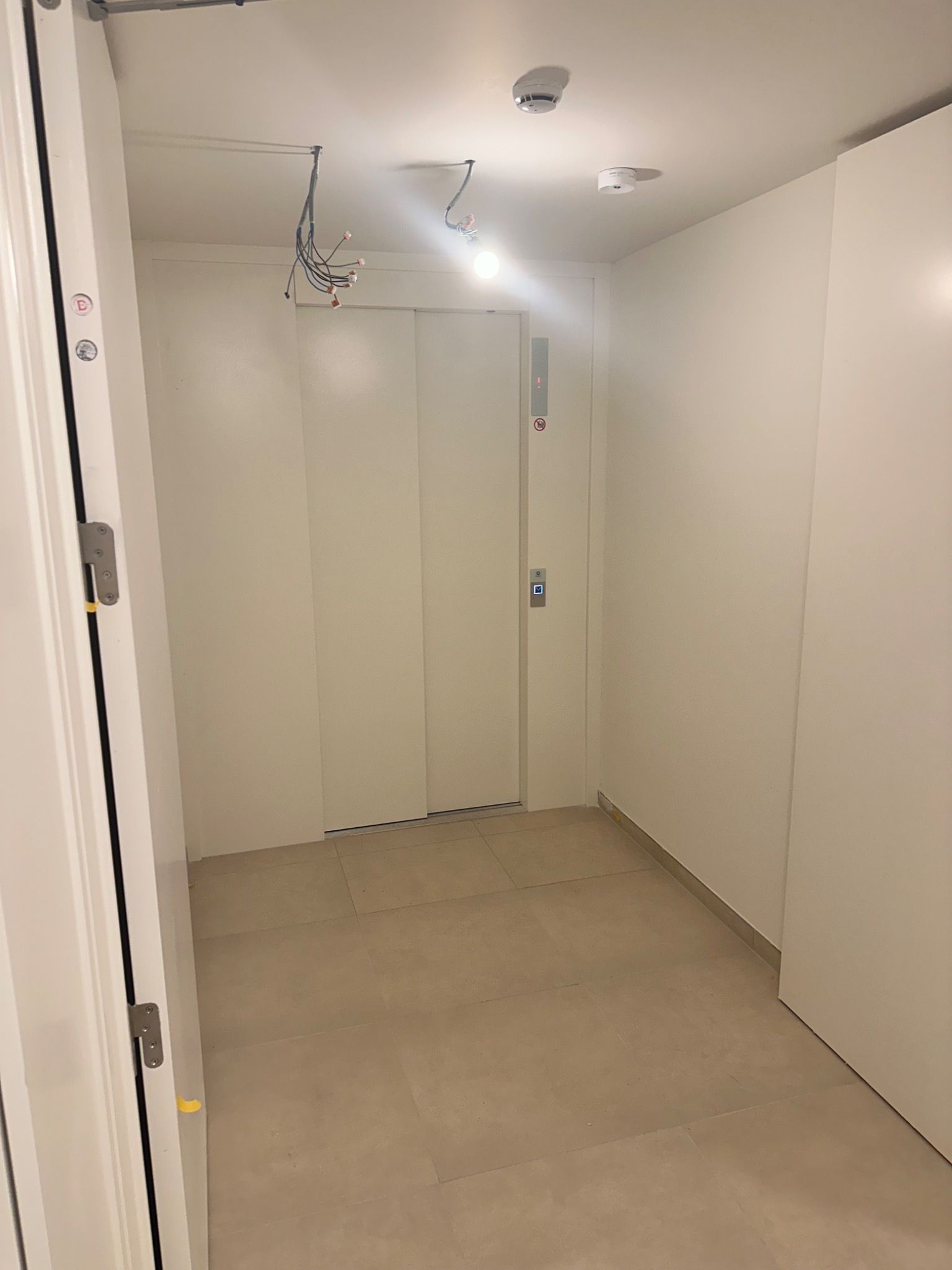 Wnętrze korytarza z białymi ścianami, jasną podłogą z płytek i białymi drzwiami. Widoczne wystające przewody elektryczne z sufitu i przycisk windy.