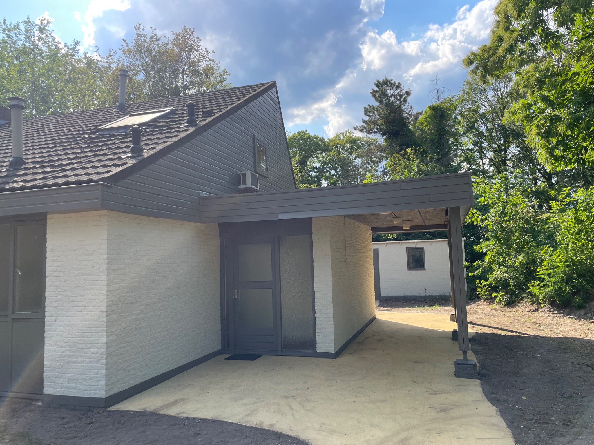 Odnowiona elewacja domu jednorodzinnego z jasną cegłą, szarą stolarką okienną i drzwiową oraz zadaszeniem nad wejściem, widoczny fragment ogrodu i błękitne niebo.