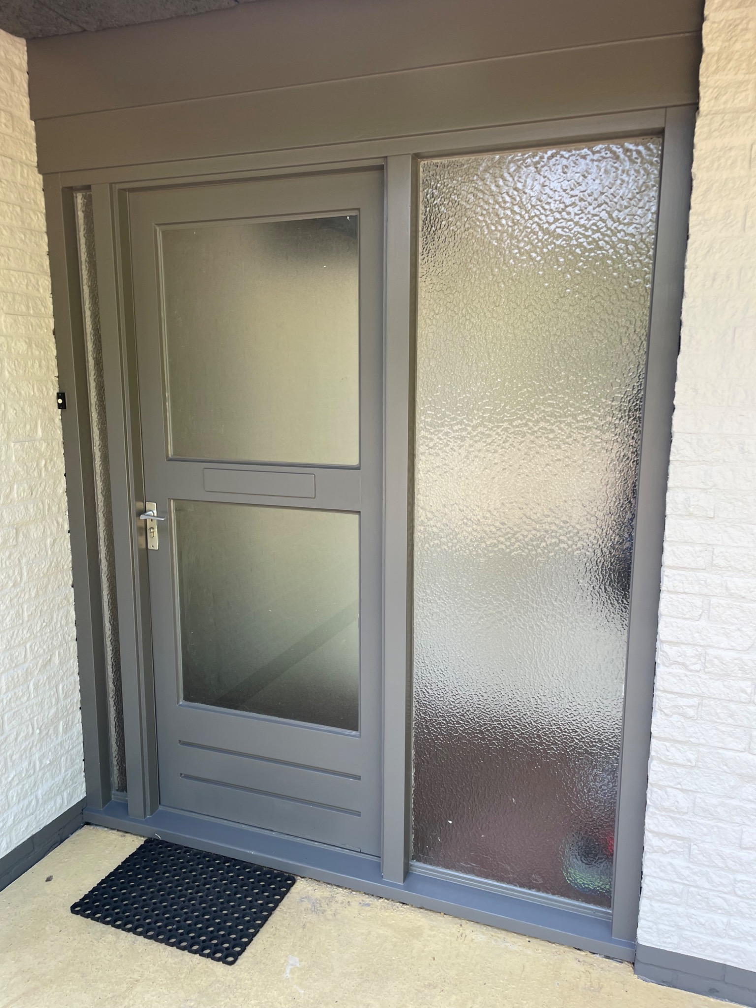 Szare drzwi wejściowe z szybą ornamentową i skrzynką na listy, zamontowane w białej ceglanej elewacji, z czarną wycieraczką u dołu.